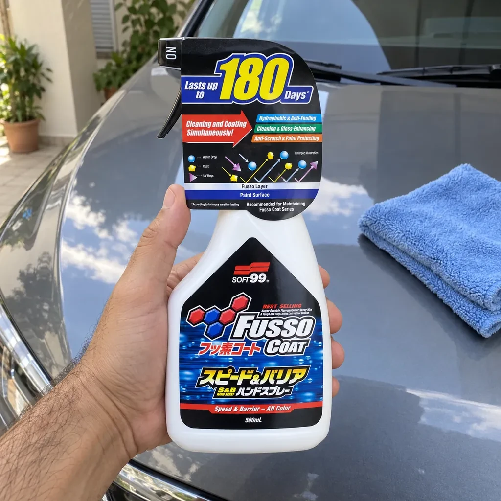 Mão segurando o frasco branco da Soft99 Fusso Coat Speed & Barrier sobre o capô cinza de um carro, com uma microfibra azul dobrada ao lado